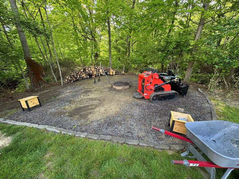 Orange track loader spreading gravel on a new patio pad around a fire ring at the edge of the woods