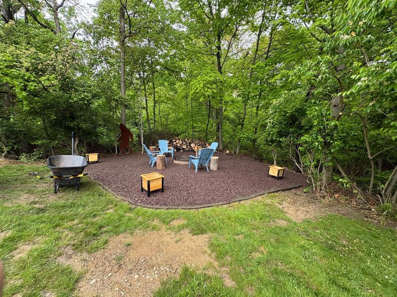 Round red gravel patio with blue chairs around a fire pit, trees behind