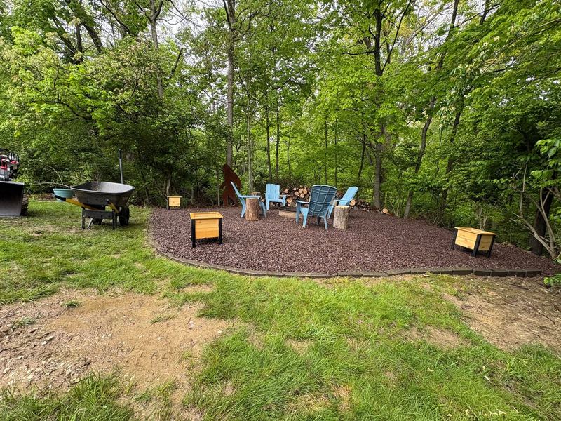 Wide view of a round red gravel fire-pit patio at the edge of a wooded backyard