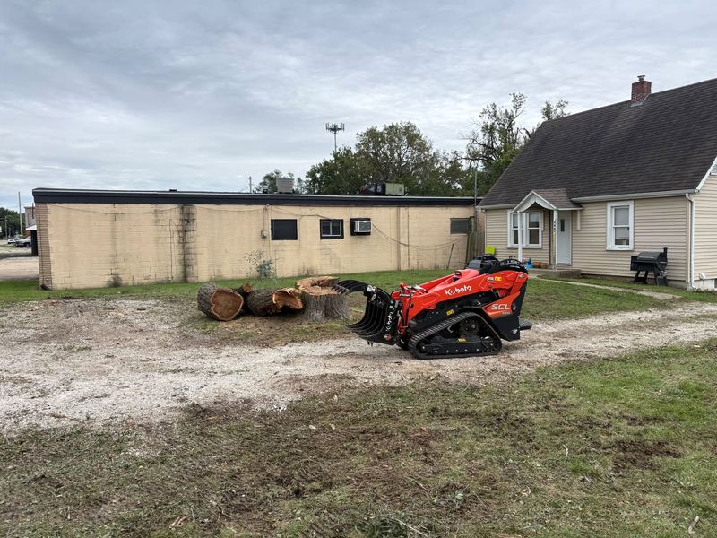 Orange Kubota track loader with a grapple next to a row of cut log rounds