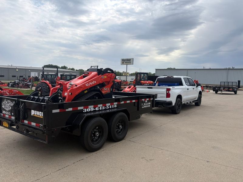 Orange Kubota track loader chained down on a black LB Tree and Land dump trailer behind a white pickup