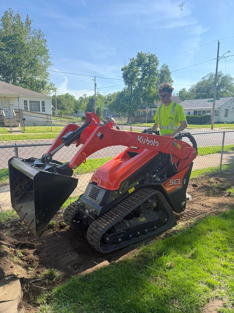 Worker on an orange Kubota track loader digging out a path along a fence